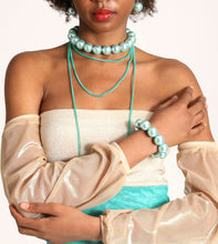 Load image into Gallery viewer, A model wearing a layered jewelry set featuring a turquoise beaded choker, a long turquoise chain necklace, a matching turquoise bracelet, and a turquoise earring, styled with a strapless cream top and sheer champagne puff sleeves.